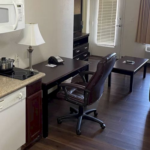 A small apartment with a kitchenette, desk, office chair, sofa, and coffee table, featuring hardwood floors and a windowed door.
