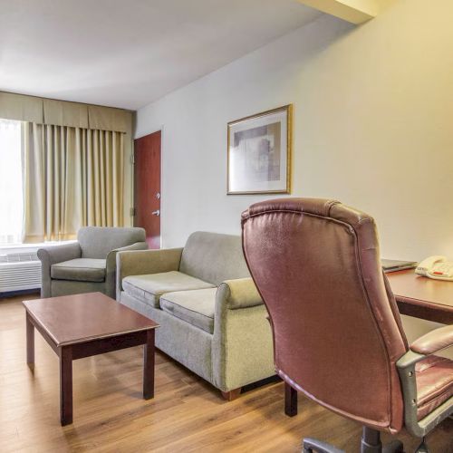 The image shows a hotel room with a desk, chair, sofa, coffee table, armchairs, and a bed near a window with curtains.