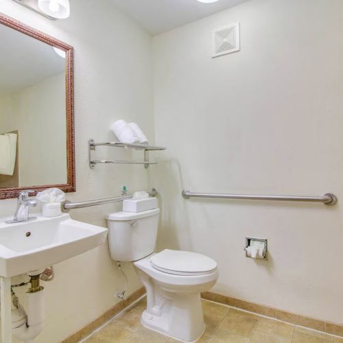 The image shows a clean bathroom with a sink, toilet, mirror, grab bars, towels on a shelf, and a wall-mounted hairdryer.