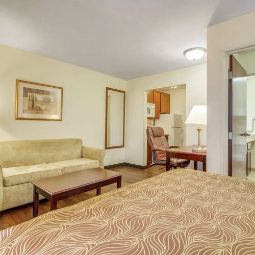 A hotel room features a bed, sofa, table, lamps, artwork, and a visible bathroom area.