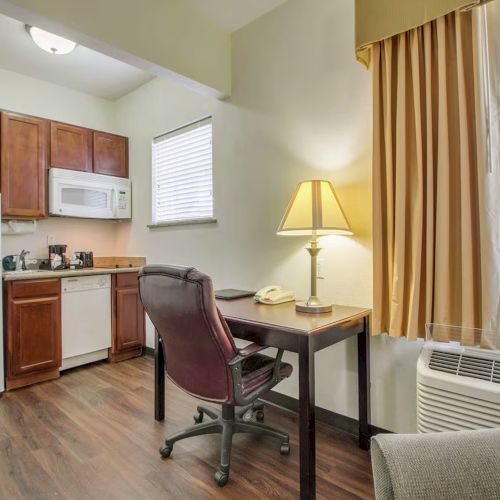 A small kitchen with fridge, microwave, and cabinets; adjacent desk with chair, lamp, phone; nearby window with curtains and air conditioner.