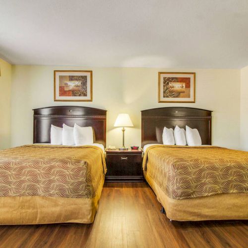 A hotel room with two double beds, wooden flooring, bedside table, lamp, artwork on the wall, and a window with curtains.
