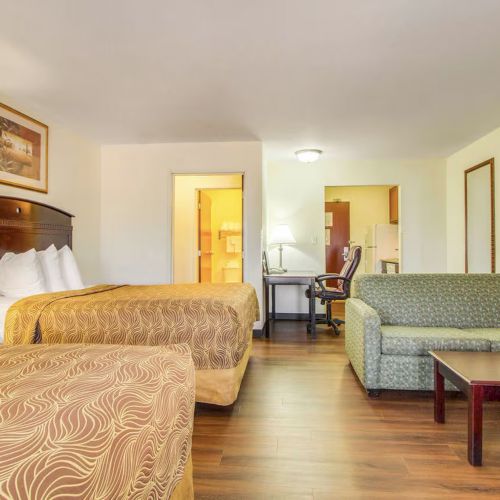A hotel room with two beds, a patterned sofa, a desk with a chair, a lamp, and wood flooring. There's an open doorway and a wall mirror.