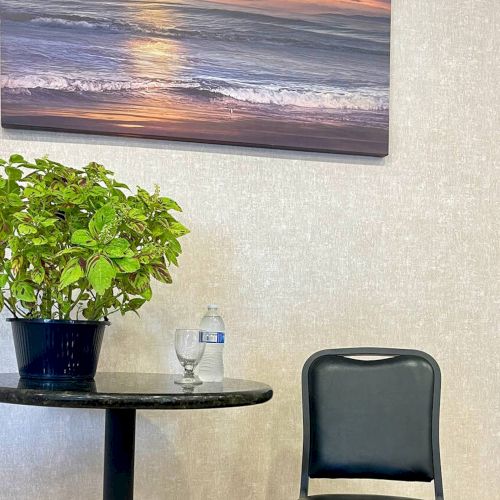 A plant on a table, a water bottle, an empty chair, and a beach sunset painting on the wall.