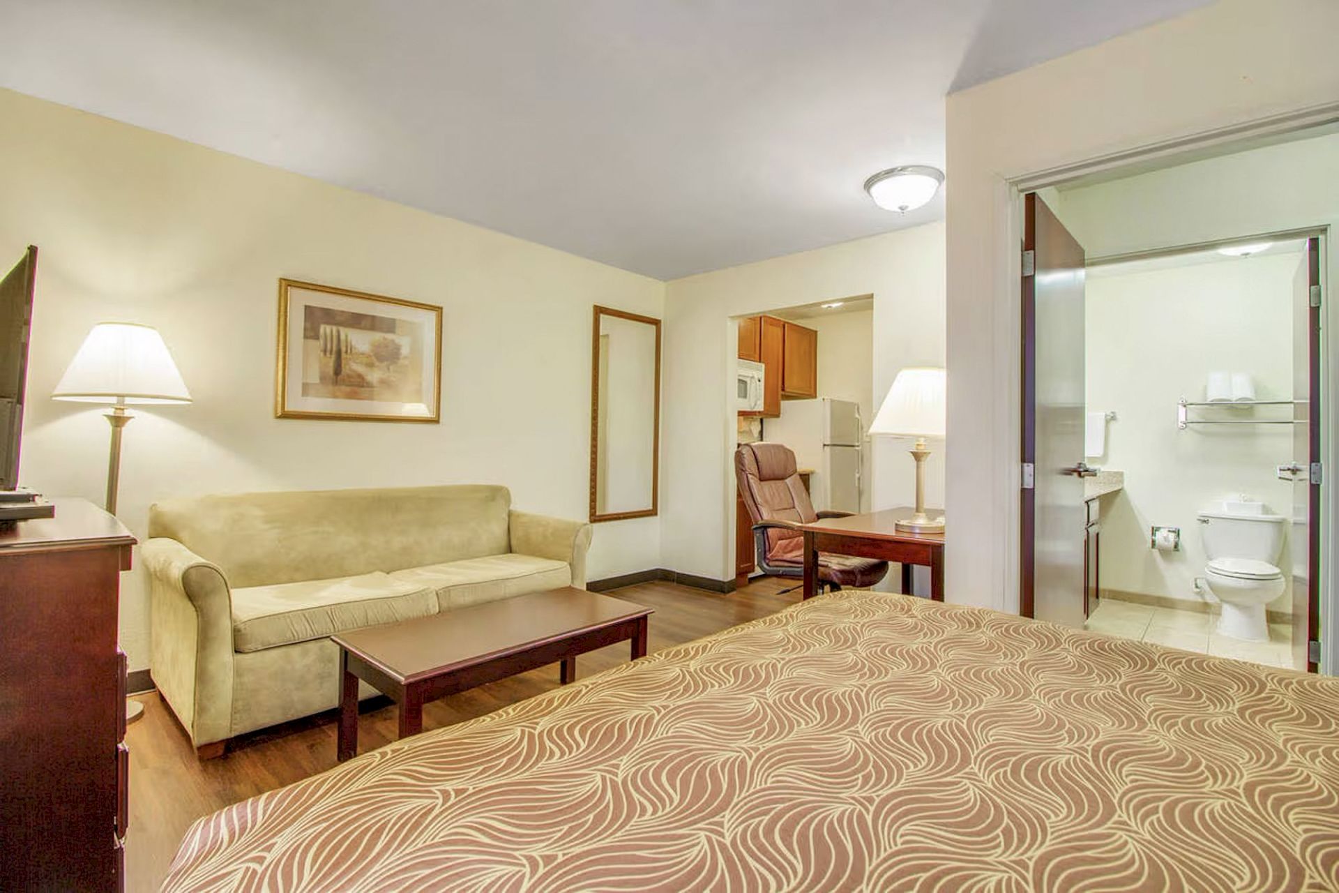 A cozy hotel room featuring a bed, sofa, coffee table, lamps, wall art, and a view into a bathroom and kitchenette area.