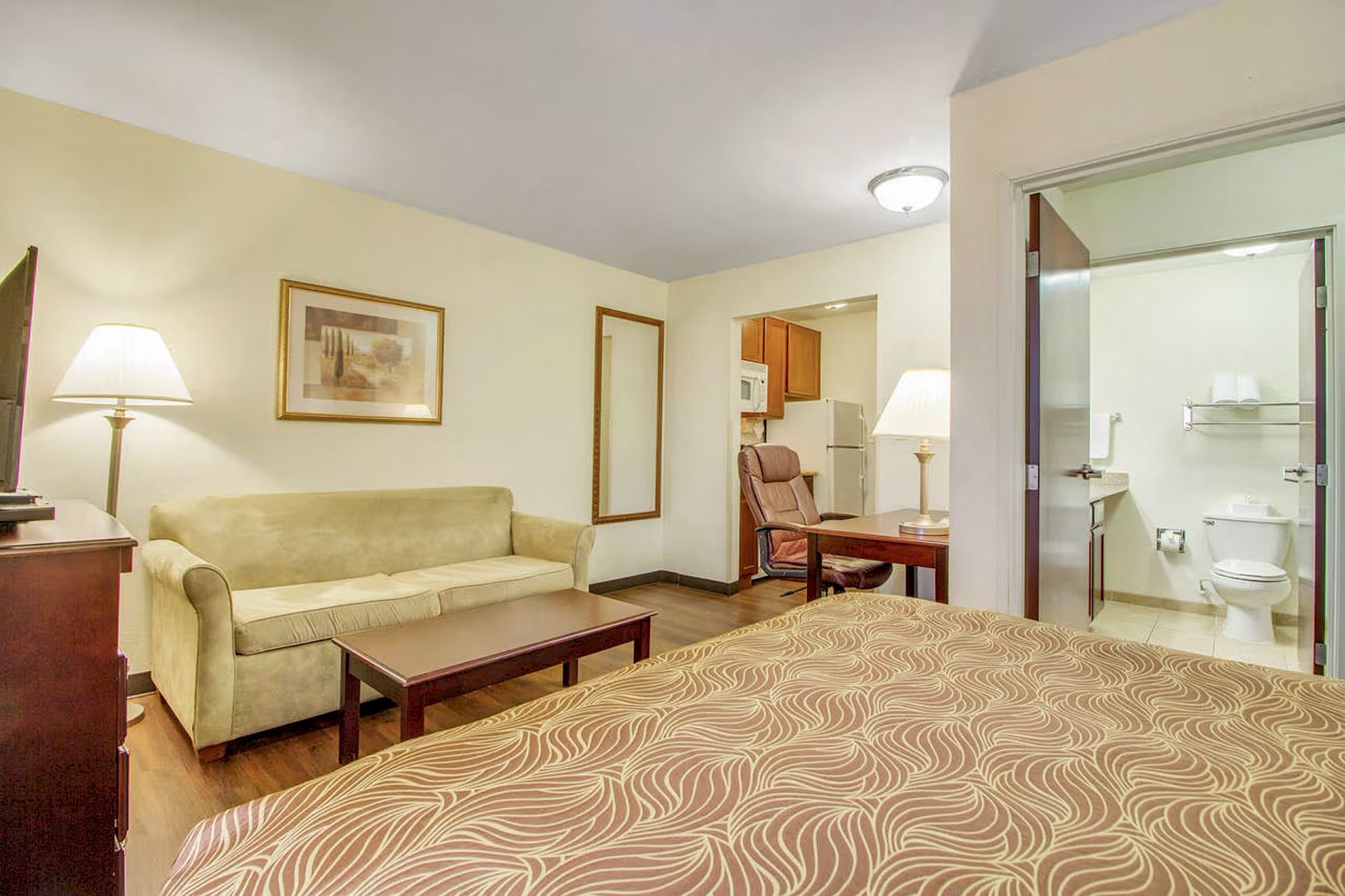 A cozy hotel room featuring a bed, sofa, coffee table, lamps, wall art, and a view into a bathroom and kitchenette area.