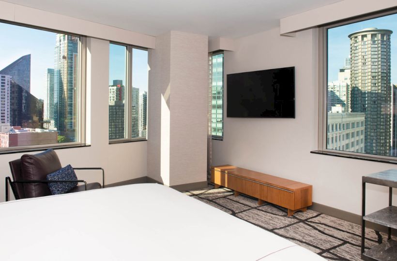 The image shows a modern hotel room with large windows, offering a city view. It features a bed, chair, wall-mounted TV, and a small table.