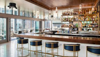 The image shows a modern bar with a sleek counter, cushioned stools, and a large selection of bottles on shelves, offering cityscape views.
