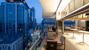 A modern rooftop terrace at dusk, with outdoor seating and tables overlooking a city skyline filled with tall buildings and lights.