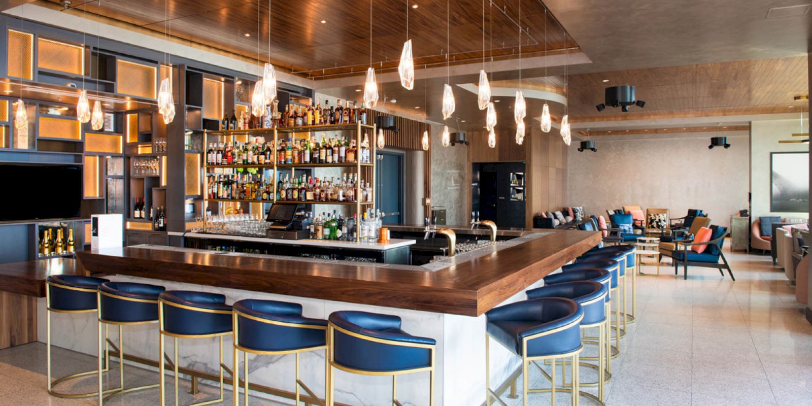 Modern bar with blue stools, wooden accents, hanging lights, and a well-stocked shelf. Cozy seating area visible in the background.