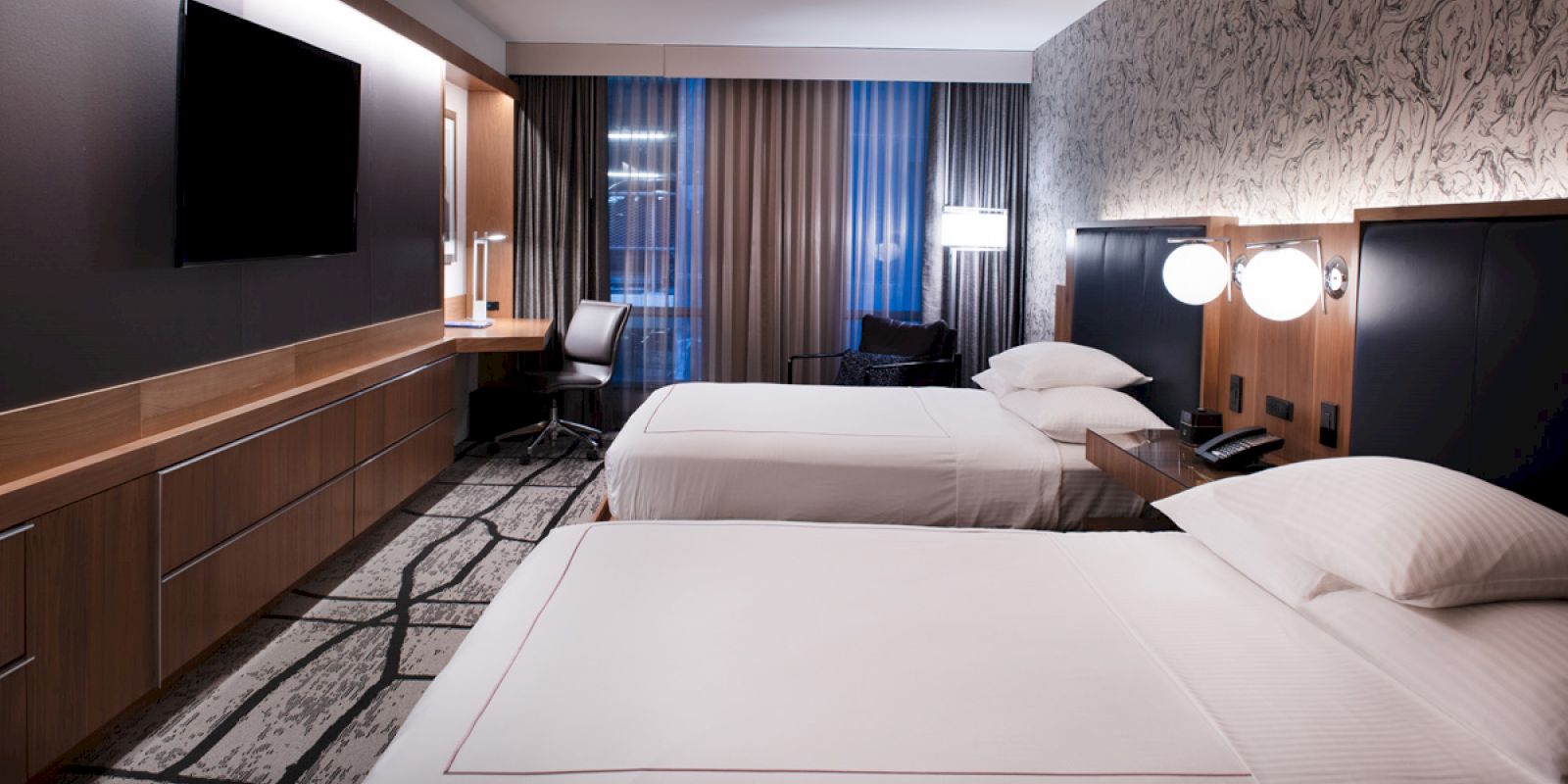 The image shows a modern hotel room with two beds, a TV, a desk, and lamps, all in a stylish, contemporary design.