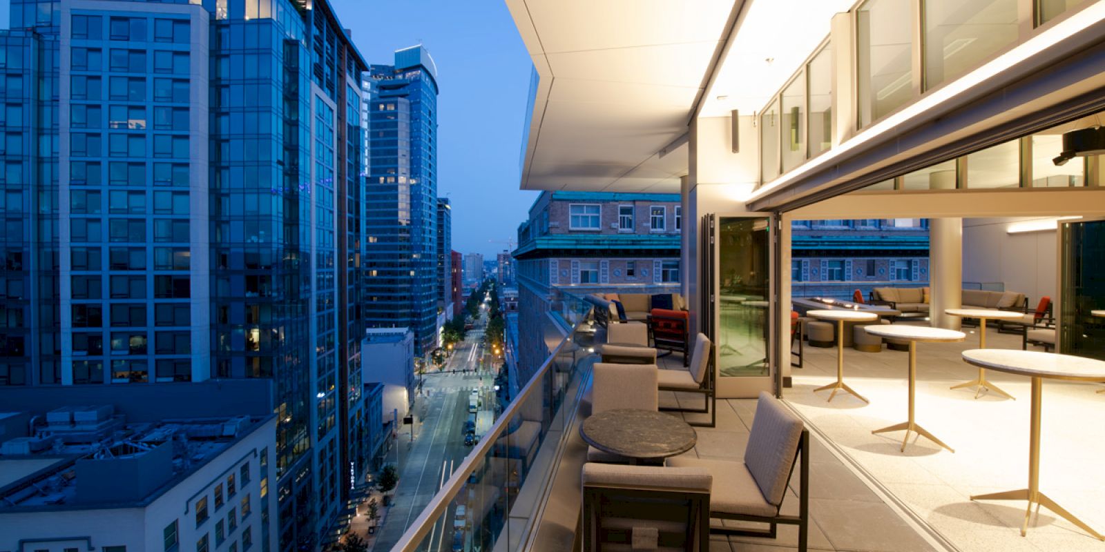 A modern cityscape view from a terrace at dusk, featuring glass skyscrapers and outdoor lounge seating, under a softly lit overhang.