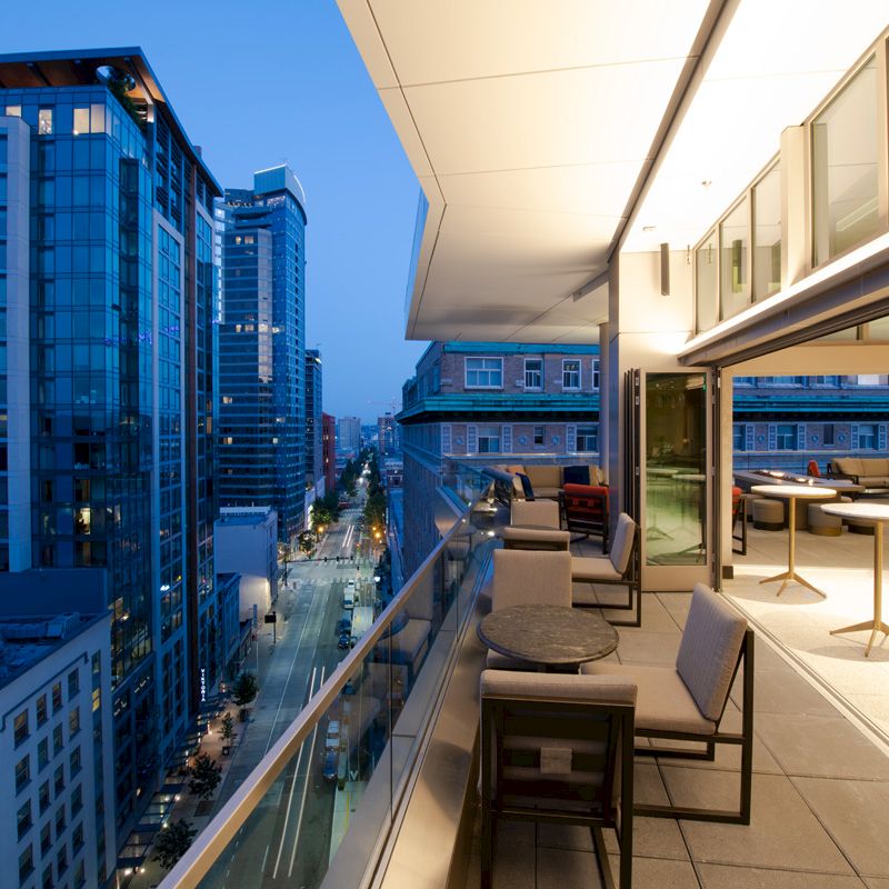 A modern rooftop terrace with seating overlooks a cityscape of tall buildings and a street, all under a twilight sky.