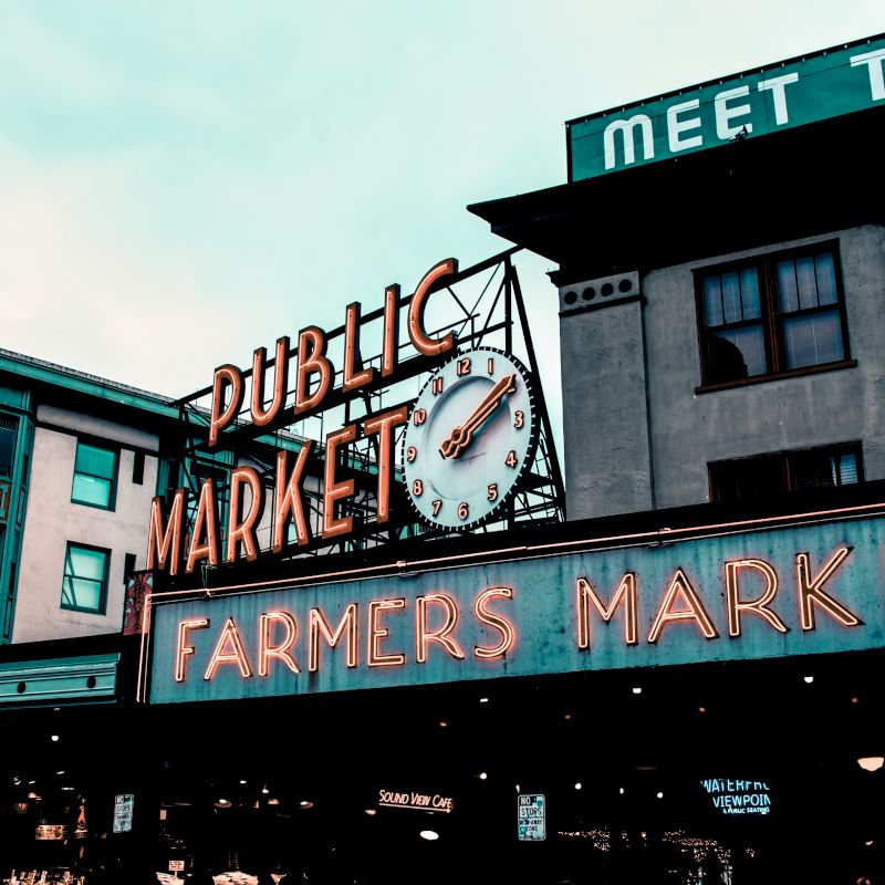 A neon sign displays "PUBLIC MARKET" and "FARMERS MARKET" with a clock, located on a building in an urban setting.