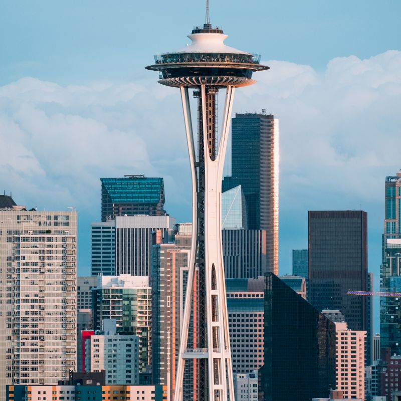 The image shows the Space Needle with a backdrop of Seattle's skyline, featuring several tall buildings under a partly cloudy sky.