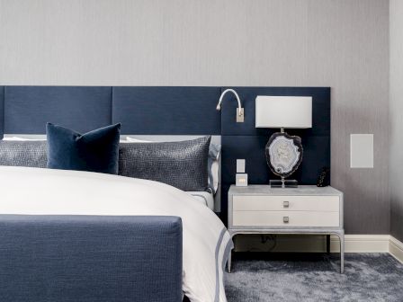 A modern bedroom with a blue and white color scheme, featuring a bed, nightstand, lamp, and decorative items on a gray carpeted floor.