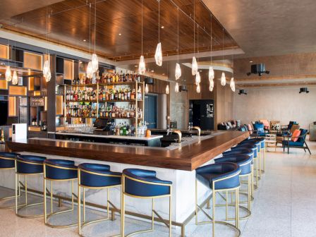 A modern bar area with a central counter, blue bar stools, pendant lights, and shelves stocked with bottles and glassware.