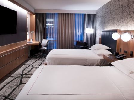 A modern hotel room with two beds, a wall-mounted TV, desk, and soft lighting. The decor is sleek with neutral tones.