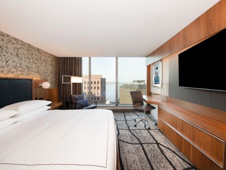 A modern hotel room with a large bed, wall-mounted TV, desk, and window view of buildings outside.