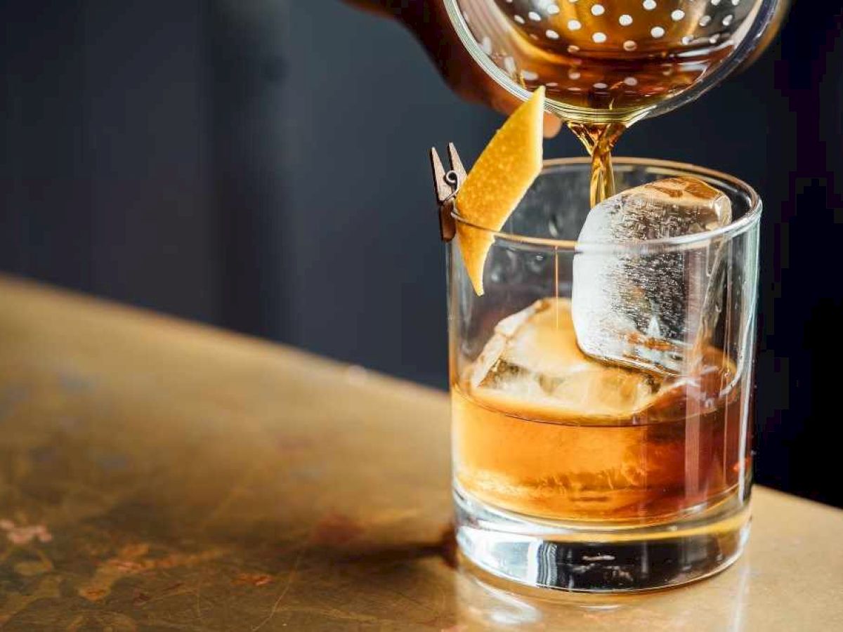 A cocktail being poured into a glass with ice and a twist of citrus peel on the rim.
