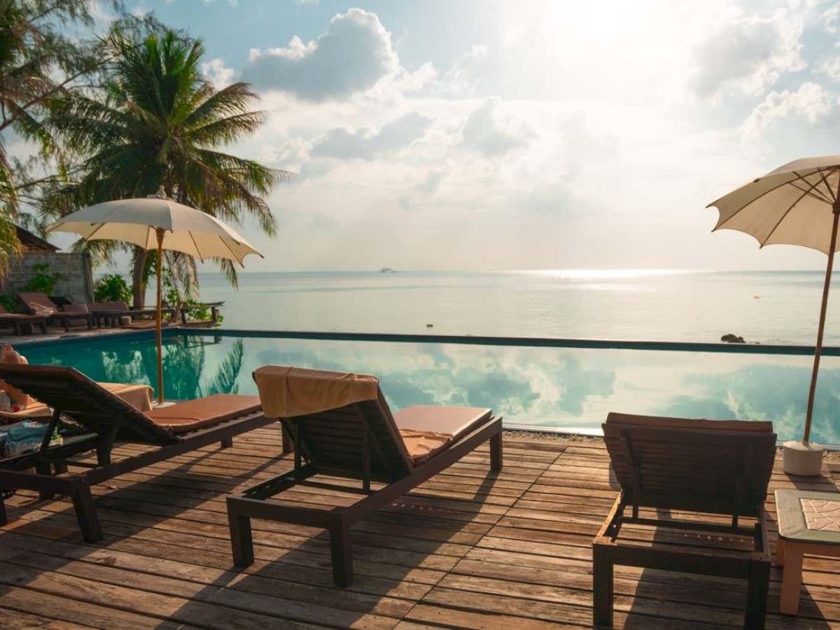Poolside view with loungers and umbrellas facing a calm sea, under a partly cloudy sky with palm trees in the background.