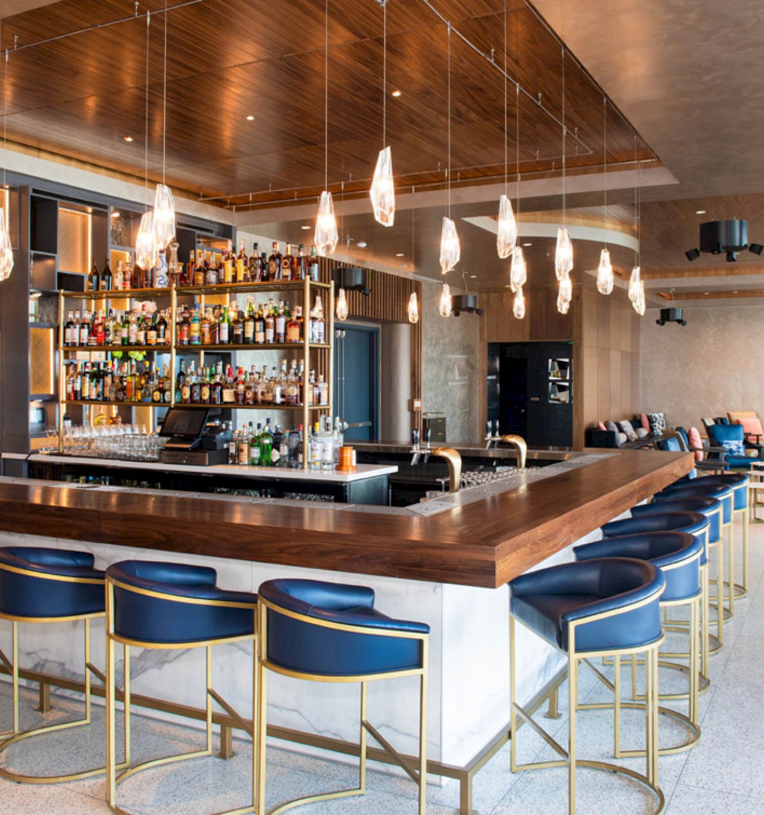 The image shows a modern bar with high stools, pendant lights, a stocked bar counter, and a spacious seating area with a contemporary design.