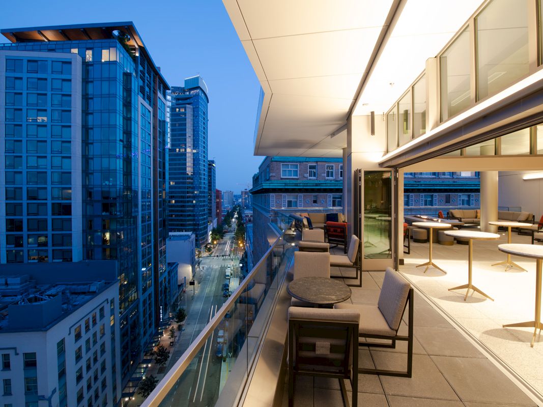 A modern cityscape with tall buildings at dusk, featuring a sleek balcony with seating, overlooking an urban street view.