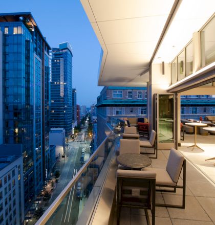 A modern terrace overlooks a cityscape with tall buildings and a lit-up street at dusk, featuring outdoor seating and tables.