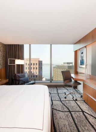 A modern hotel room with a large bed, wall-mounted TV, desk, and window offering a city view.