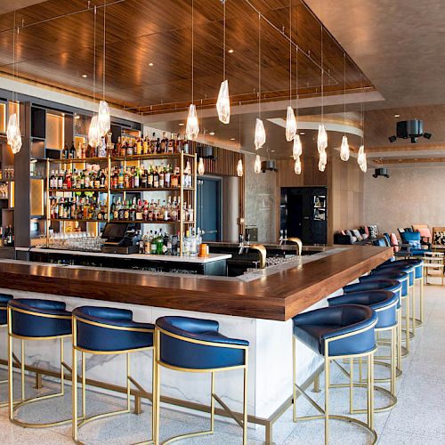 A modern bar with a central counter, blue bar stools, hanging lights, and a well-stocked shelf, located in a stylish lounge area.