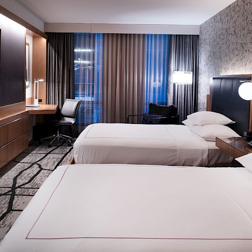 A hotel room with two beds, a TV, desk, lamp, and curtains. The design is modern with a neutral color palette and textured wall.