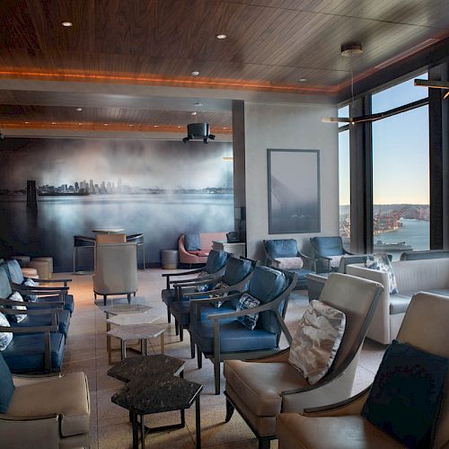 A modern lounge area with stylish seating, a city mural, and large windows showcasing a scenic view of the water and sky.