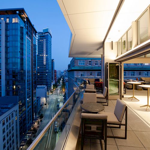 A modern balcony overlooking a cityscape with illuminated buildings and street, featuring seating and tables.