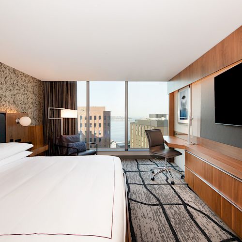 A modern hotel room with a bed, large TV, desk, and a window offering a city view.