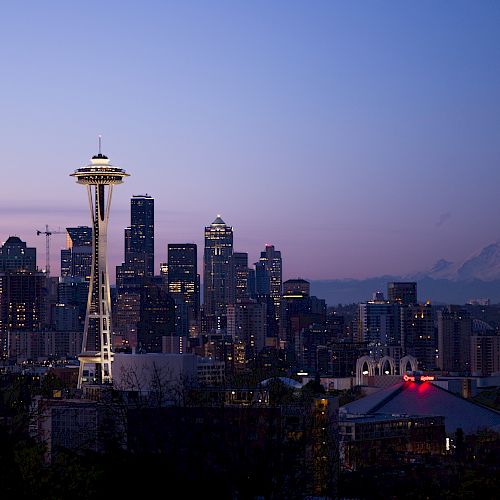 The image shows the Seattle skyline at dusk, featuring the Space Needle, with Mount Rainier visible in the background under a clear sky.