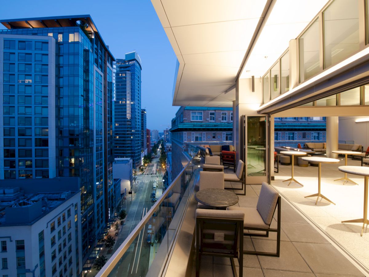 An urban rooftop terrace at dusk with modern furniture, overlooking a cityscape of tall glass buildings under a clear evening sky.