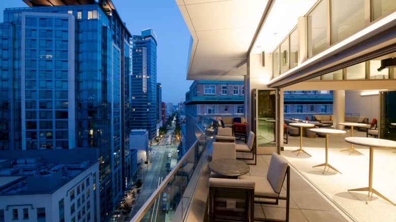 An urban rooftop terrace at dusk with modern furniture, overlooking a cityscape of tall glass buildings under a clear evening sky.