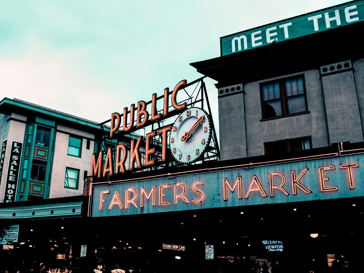 Image of a public market with a large clock and neon sign reading "FARMERS MARKET." Buildings and signage are visible in the background.