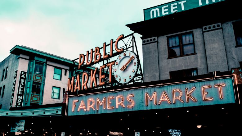 Image of a public market with a large clock and neon sign reading "FARMERS MARKET." Buildings and signage are visible in the background.