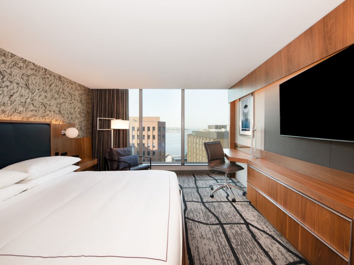 A modern hotel room with a large bed, a TV, a desk by the window, and a city view in the background.