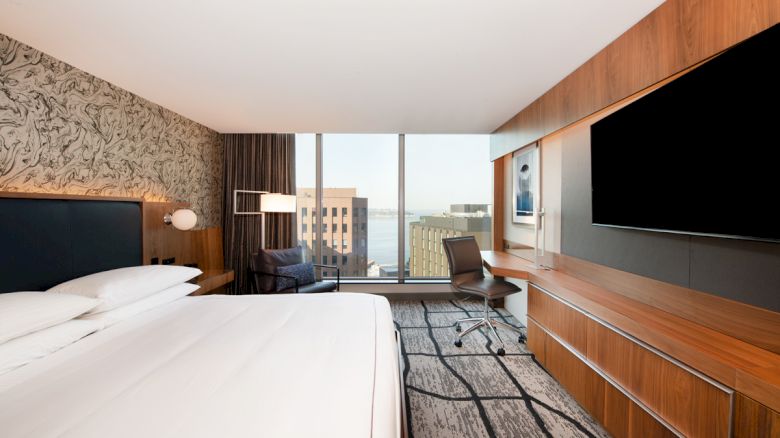 A modern hotel room with a large bed, a TV, a desk by the window, and a city view in the background.