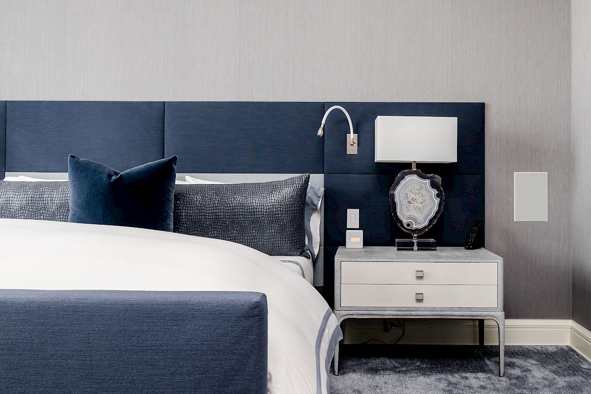 A modern bedroom with a navy and gray theme, featuring a bed, nightstand, lamp, and decorative pillows.