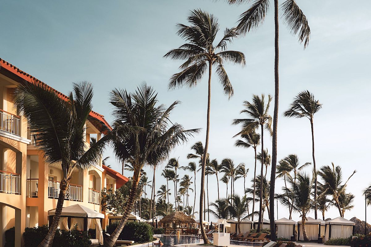 A tropical resort scene with a pool, lounge chairs, palm trees, and a building in the background under a clear sky.