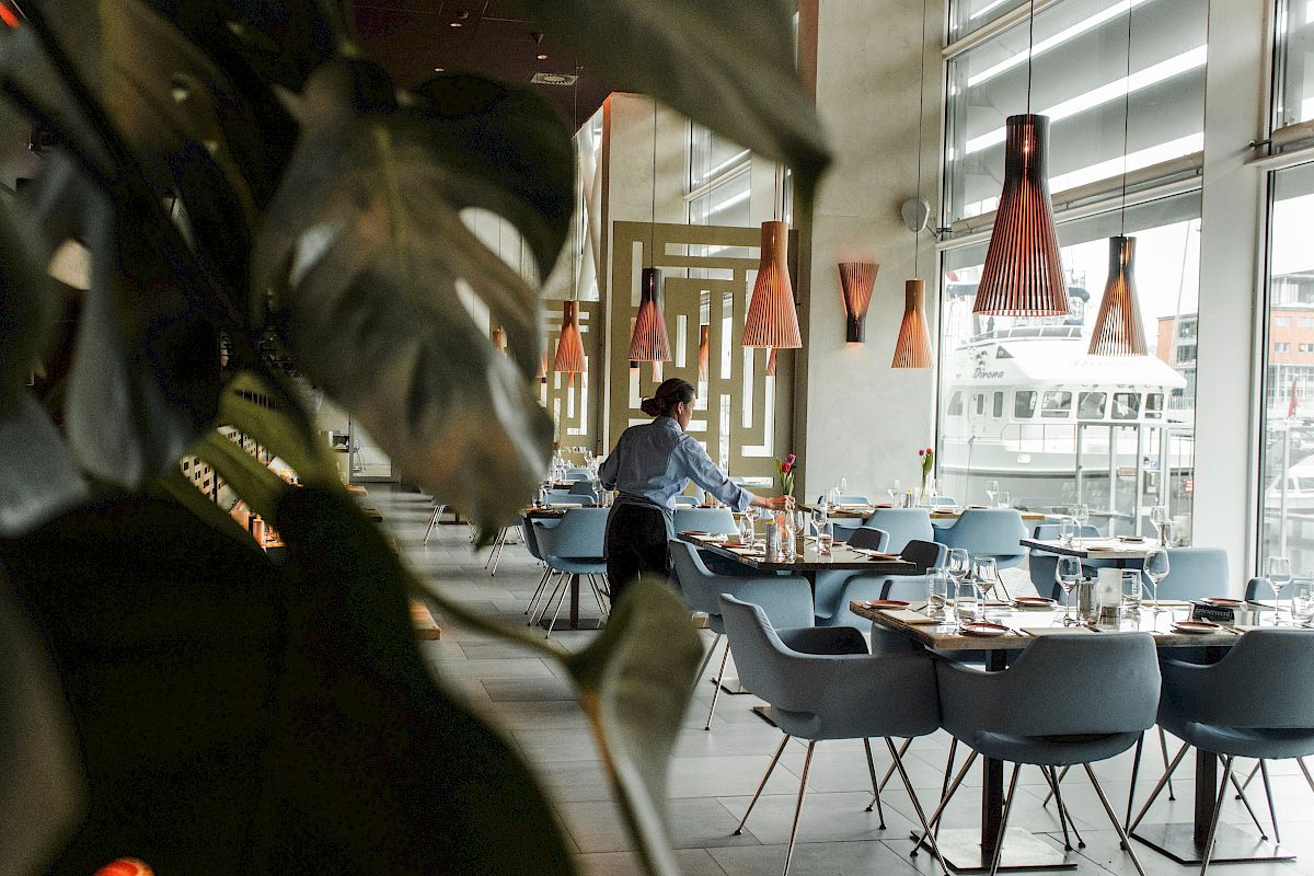 A person sets tables in a modern restaurant with stylish lighting and large windows, partially obscured by a plant in the foreground.