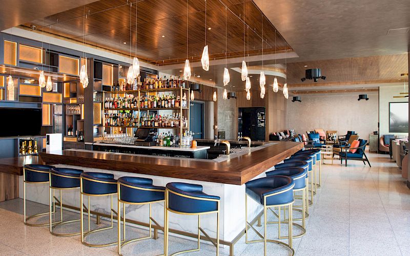 The image shows a modern bar with a wooden counter, blue stools, hanging lights, and a well-stocked display of bottles.