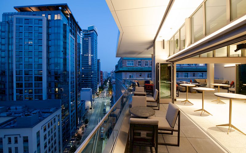 A modern rooftop terrace with seating overlooks an urban street and skyscrapers at dusk, featuring sleek design and ambient lighting.
