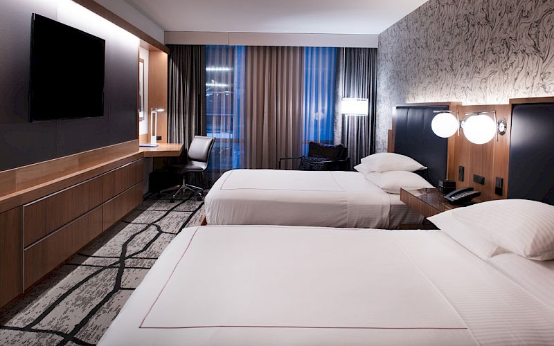 The image shows a hotel room with two neatly made beds, a wall-mounted TV, a desk, chair, lamps, and a window with curtains.